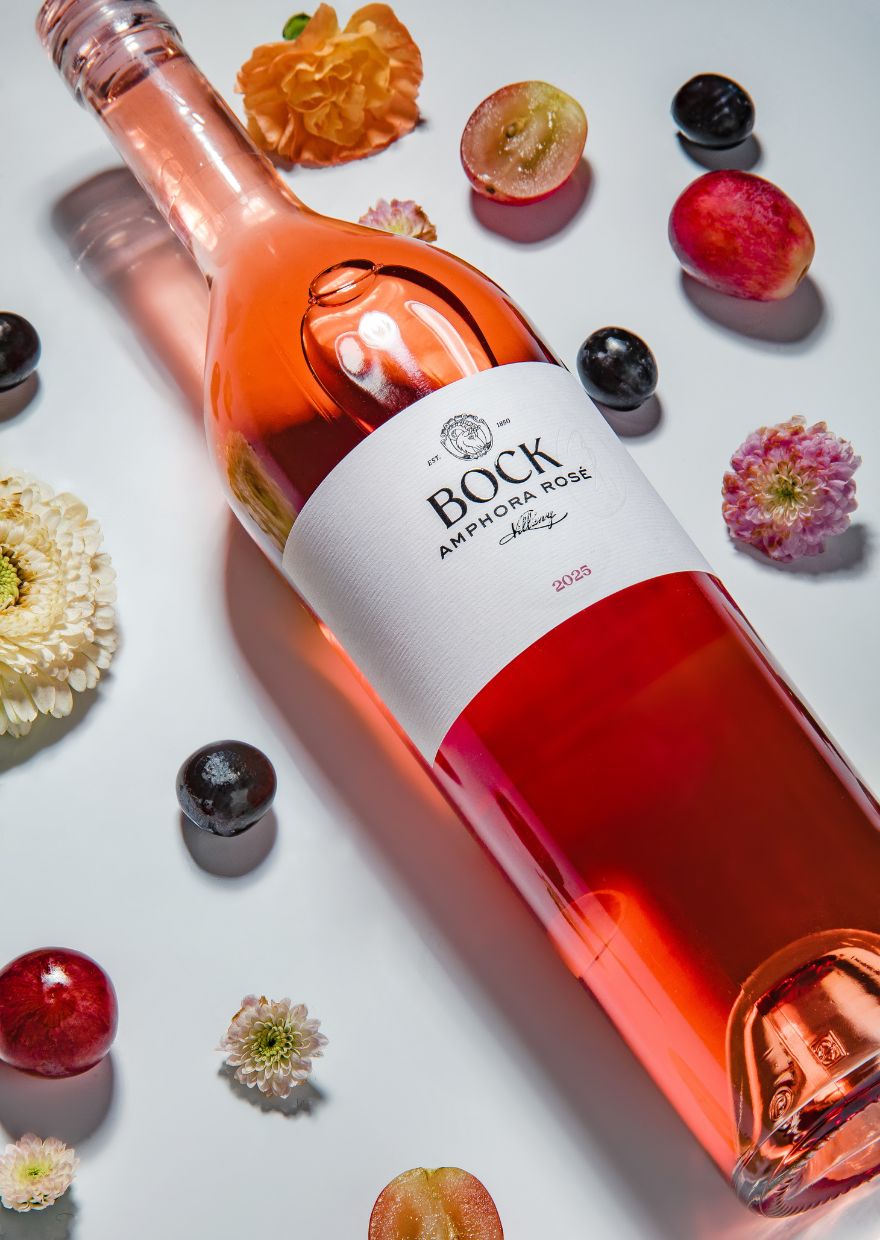 Bock Amphora Rosé 2025 Bottle of Bock Amphora Rosé wine on a white surface, surrounded by flowers and small fruit like grapes and peaches/plums.
